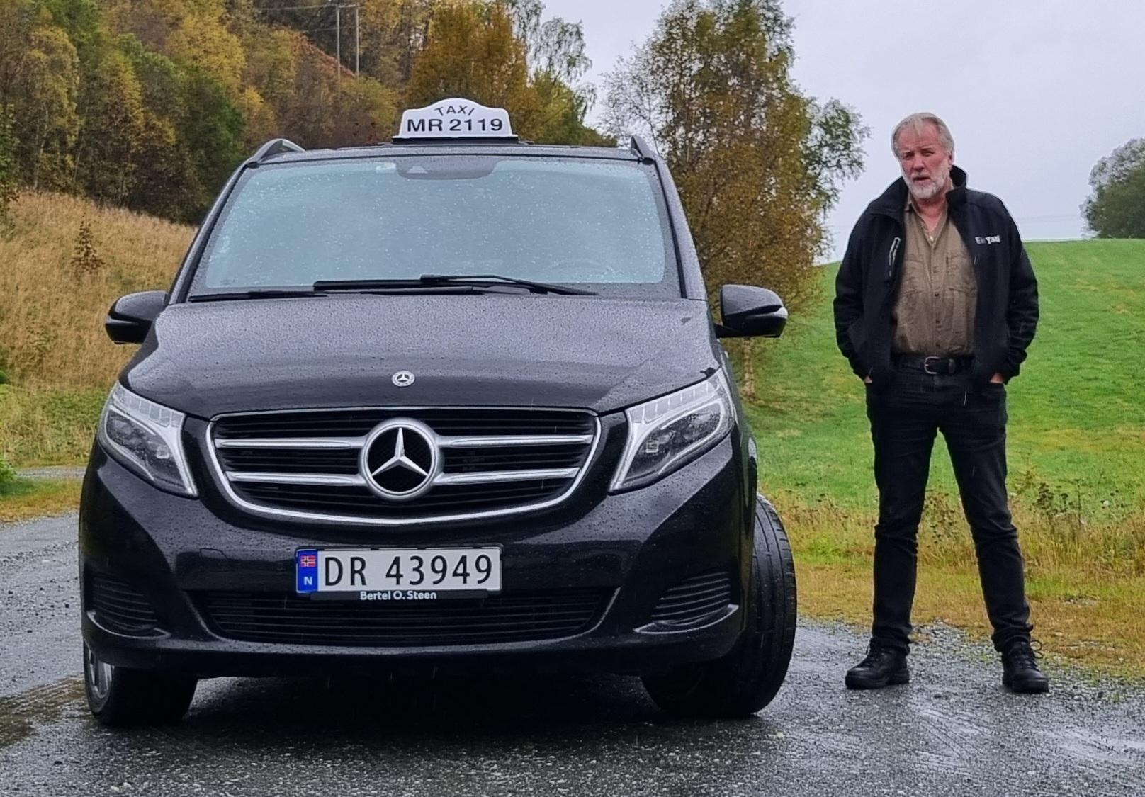 En mann står ved siden av en svart Mercedes taxi på en regnfull vei med høstfarger i bakgrunnen.