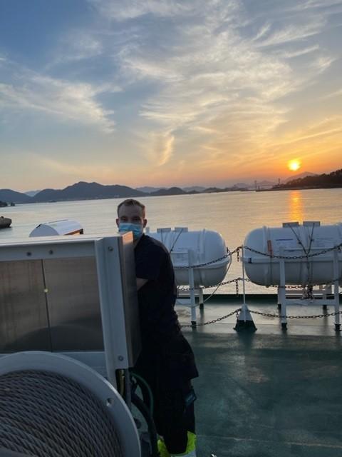 En mann i arbeidsklær står ved en livbåt på en båt med solnedgang i bakgrunnen.