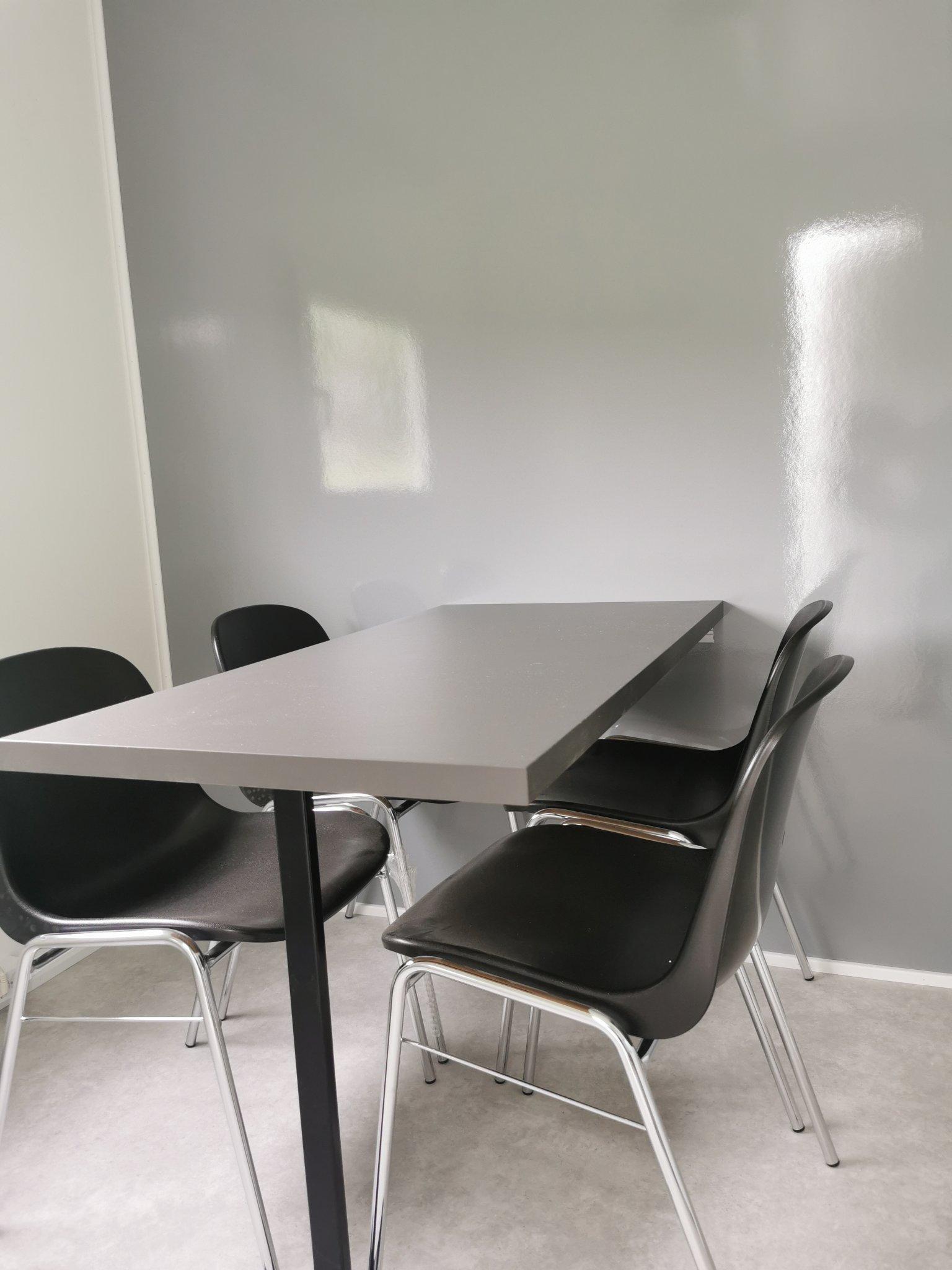 Et moderne rom med et grått bord og svarte stoler mot en lys grå vegg.