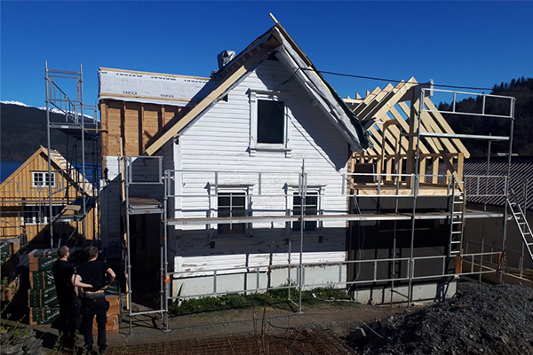 Hus under bygging med stillas, takstoler og arbeidere i solfylt landskap ved vannet.