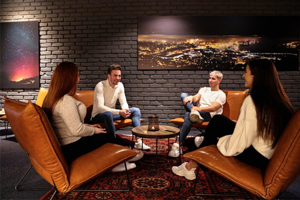 Fire unge mennesker sitter i en koselig stue med mørke vegger og moderne møbler, og diskuterer sammen.
