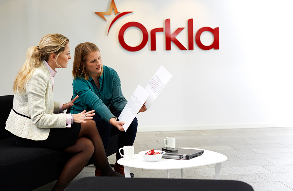 To kvinner diskuterer dokumenter i et moderne kontorlandskap med Orkla-logoen i bakgrunnen.
