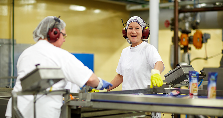 To kvinner i hvite arbeidsklær og hørselvern smiler mens de jobber på en fabrikk.