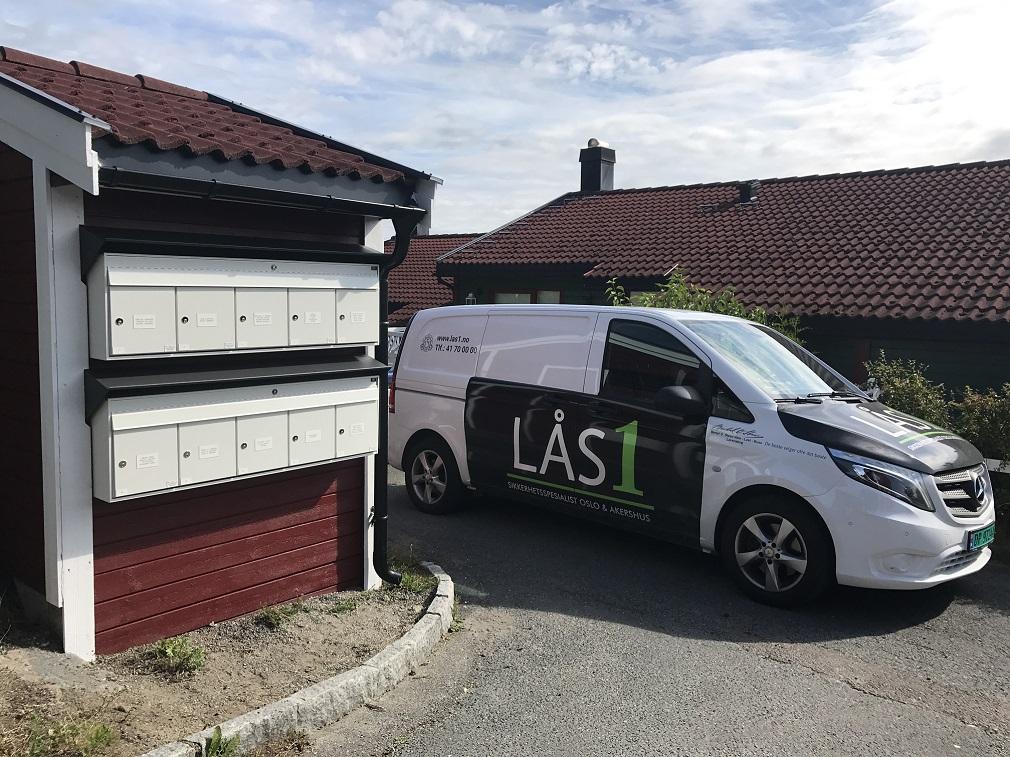 En hvit varebil med påskrift står parkert ved en postkassevegg i et boligområde.