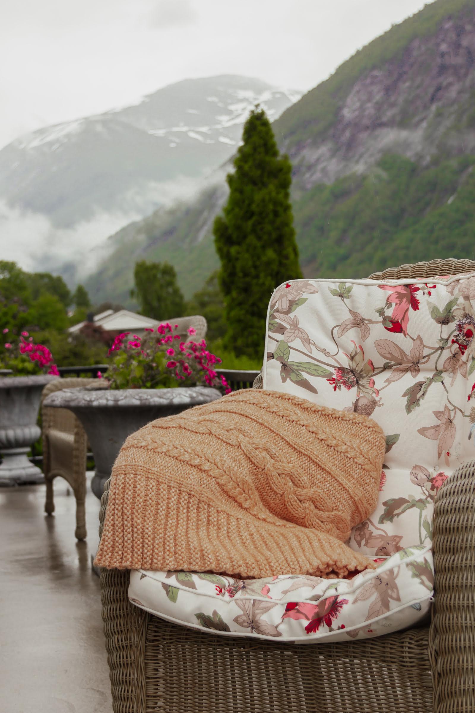En koselig stol med blomstrete pute og en oransje strikket teppe, med fjell og blomster i bakgrunnen.
