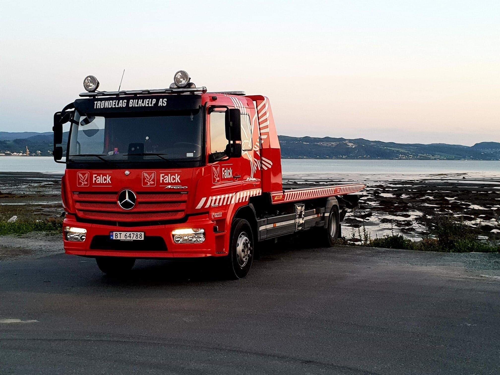 En rød bergingsbil fra Mercedes står parkert ved kysten med havet i bakgrunnen.