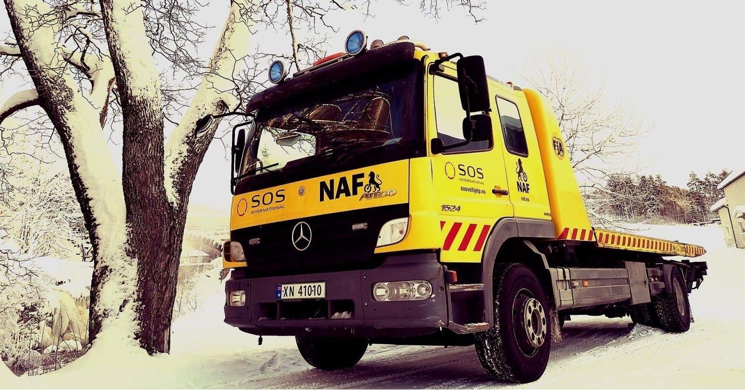 En gul bergingsbil fra NAF står parkert ved et tre i snødekt landskap.
