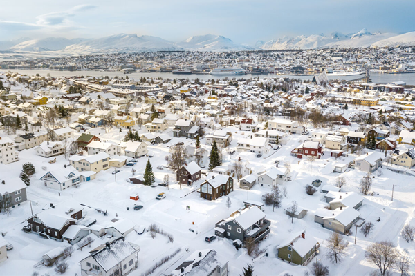 Vinterlandskap med snødekte hus og fjell i bakgrunnen, sett fra luften i en norsk by.
