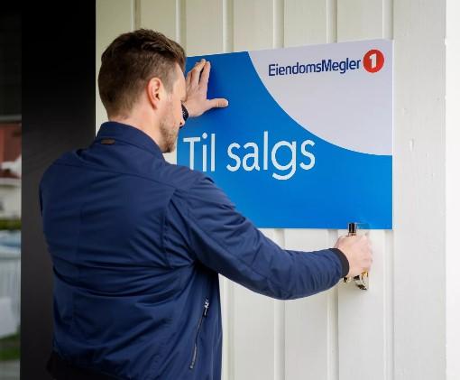 En eiendomsmegler fester et "Til salgs"-skilt på en hvit vegg.