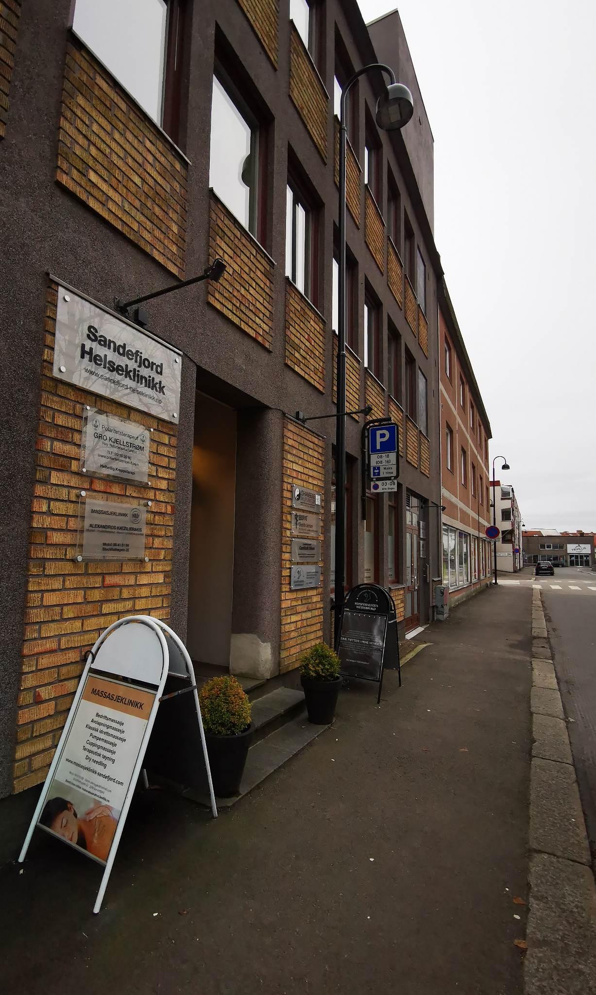 Fasade av en bygning med murstein, skilt for helsepraksis og parkeringsplass langs gaten.