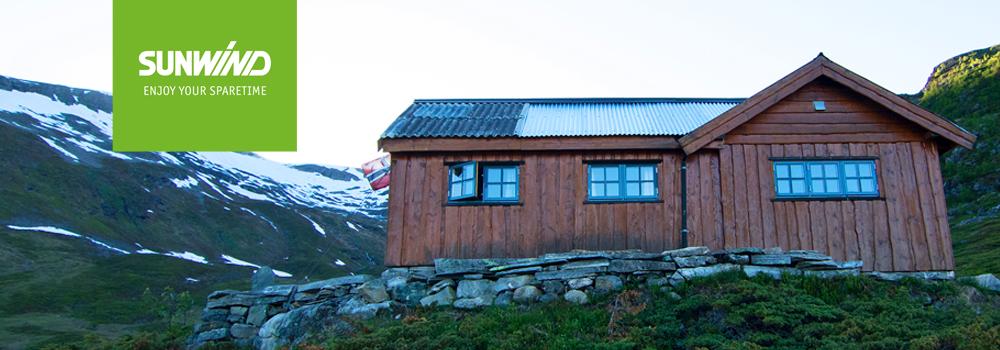 Hytte i fjellet med trevegger og vinduer, omgitt av snødekte fjell og grønt landskap.