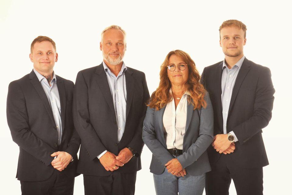 Fire profesjonelle personer i dress står sammen foran en lys bakgrunn.