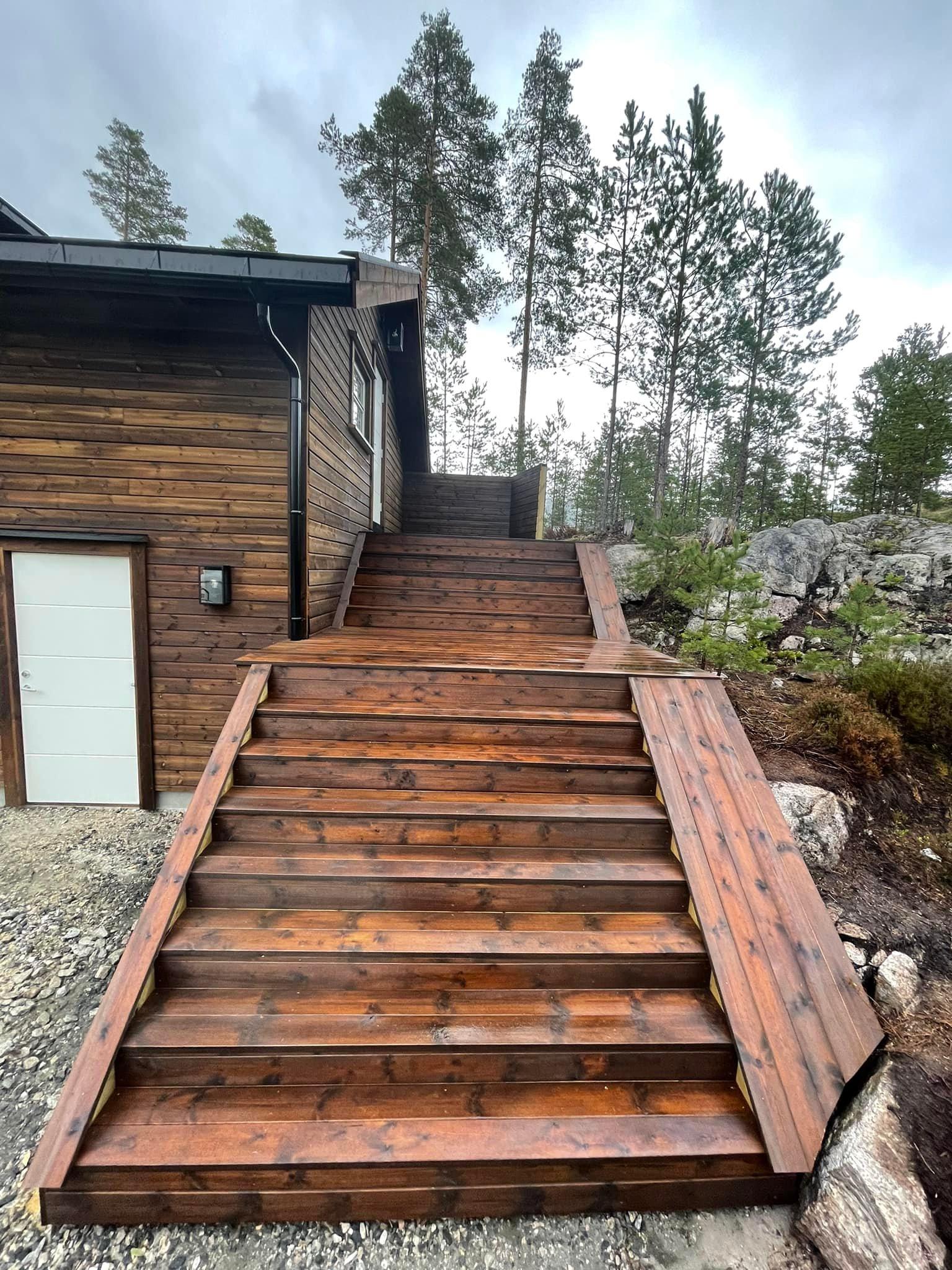 En tretrapp med flere trinn som fører opp til en hytte, omringet av trær og stein.