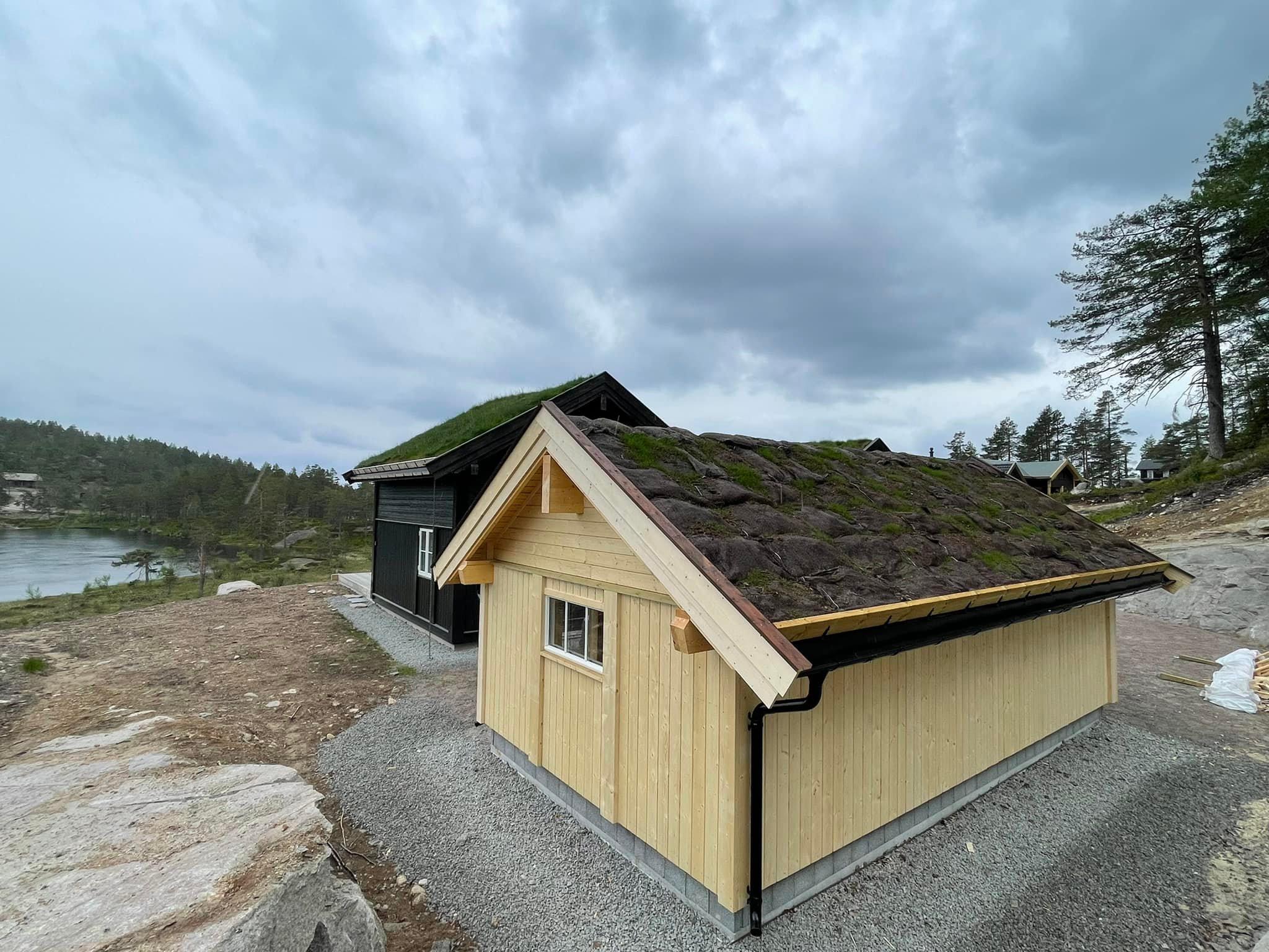 Hus med torvtak og grå steinbelagt gårdsplass, omgitt av trær og en innsjø i bakgrunnen.