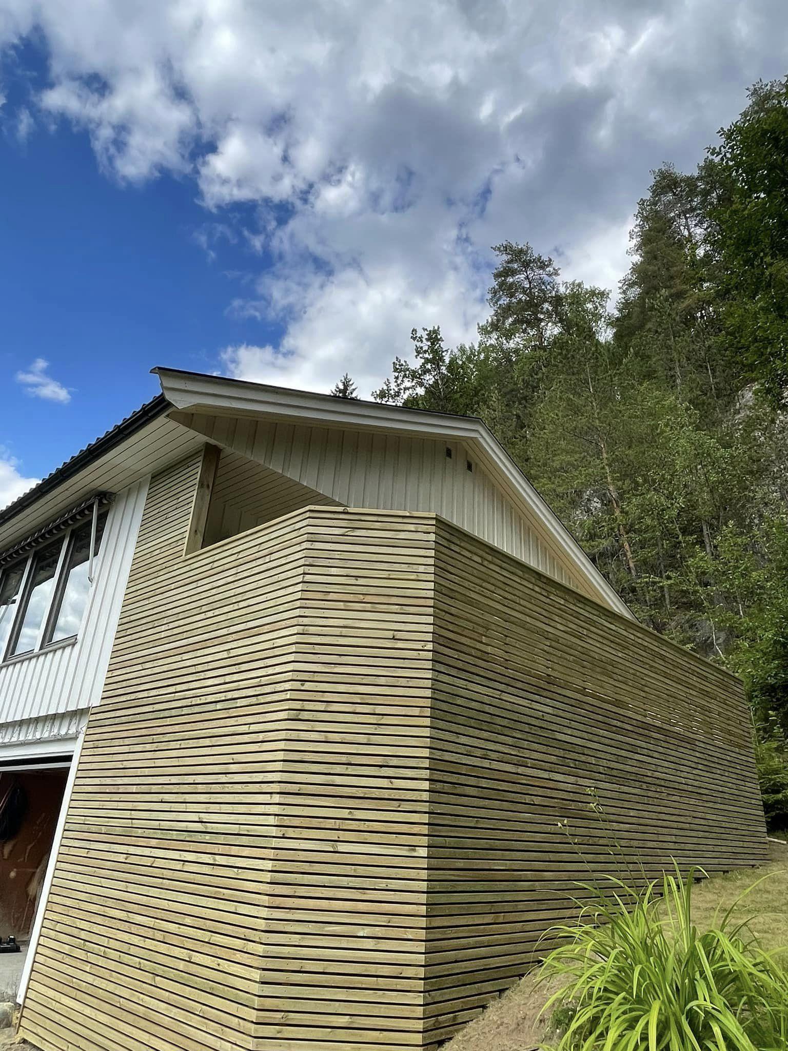 Moderne hus med en bølgete trevegg, omgitt av grønn vegetasjon og en blå himmel med skyer.