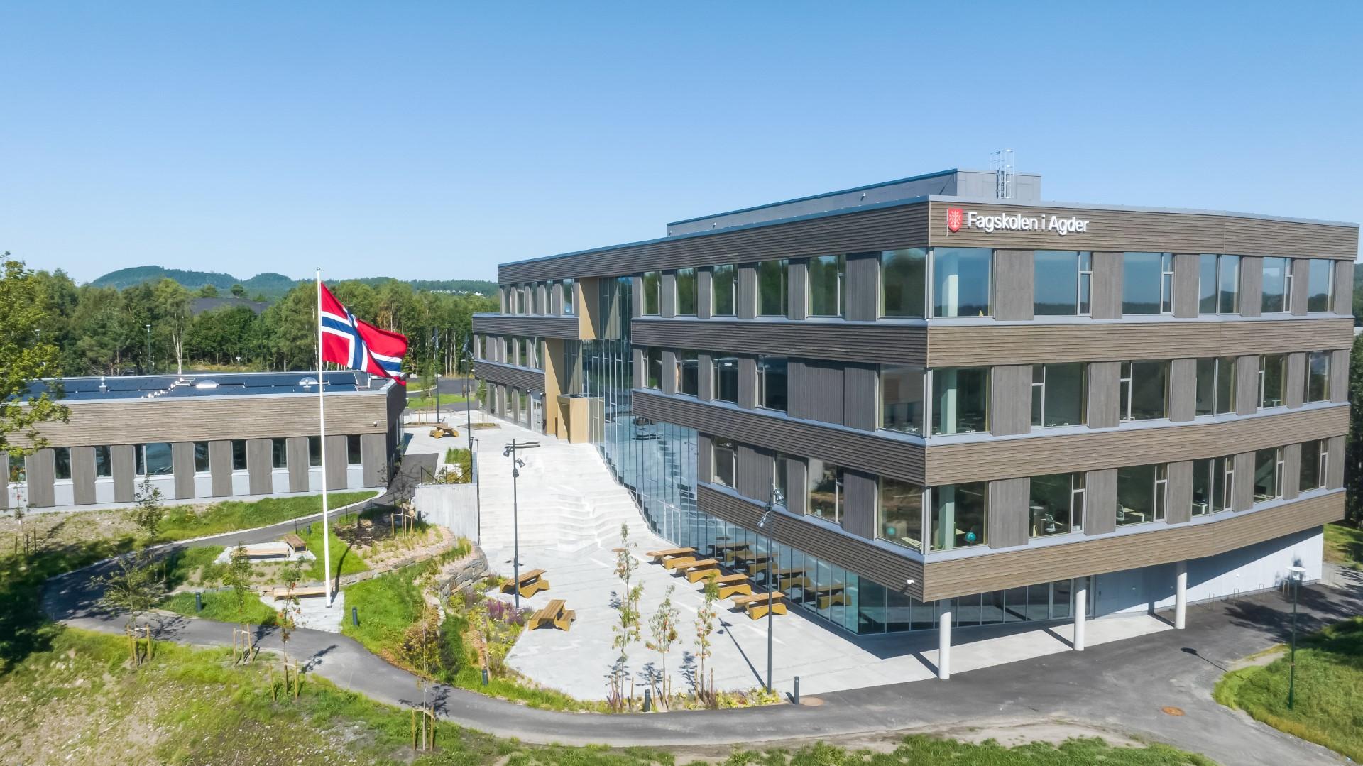 Moderne kontorbygg med norsk flagg, grønt område og solfylt himmel i bakgrunnen.