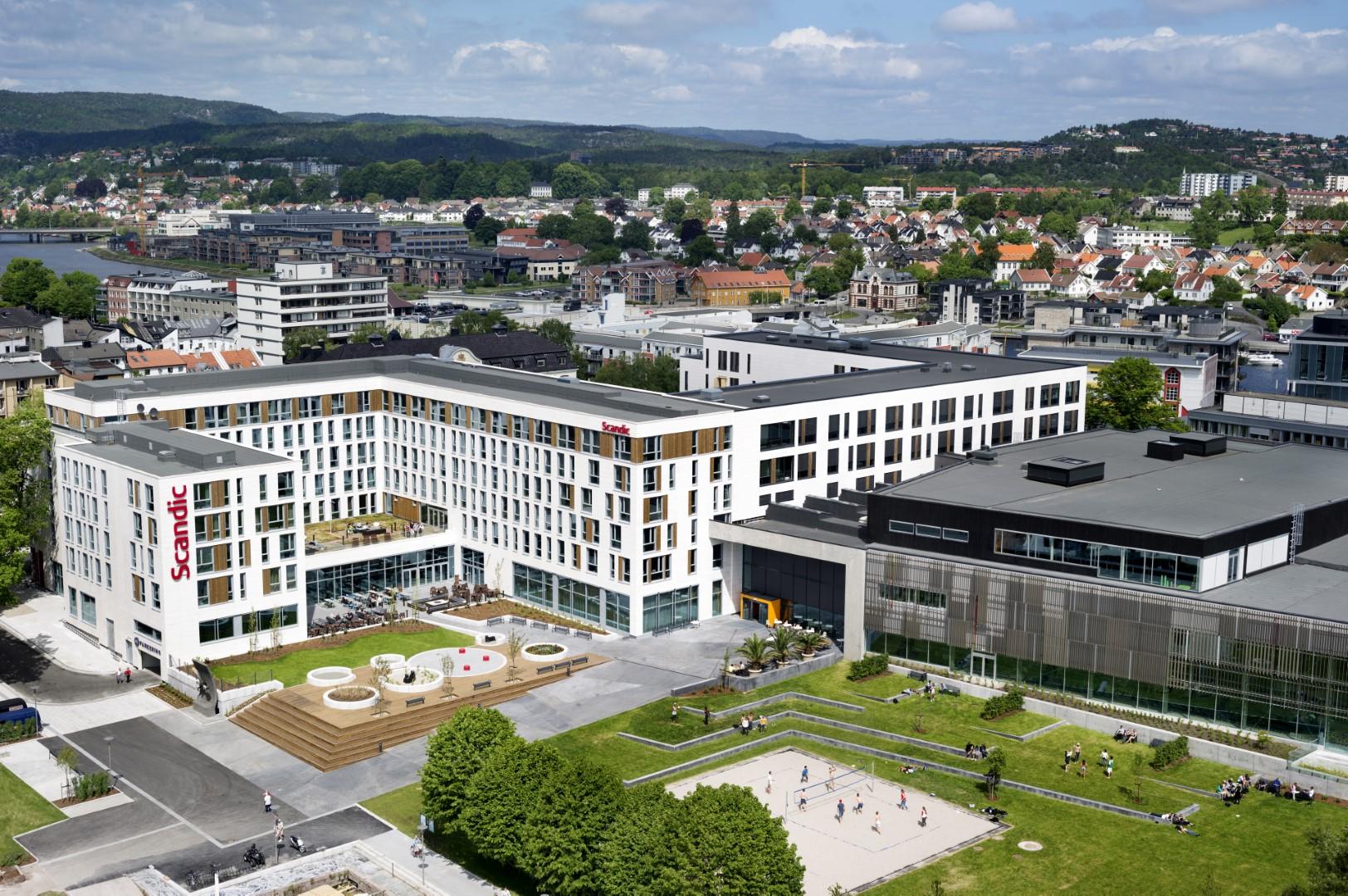 Moderne kontorbygg med grønne områder og lekeplass, omgitt av bylandskap og blå himmel.