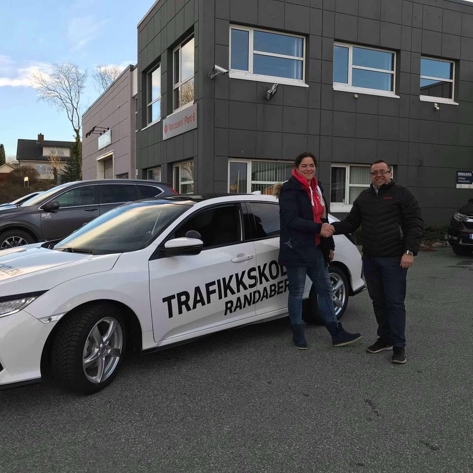 To personer står ved en bil med teksten "Trafikkskole Randaberg" på siden, foran en bygning.