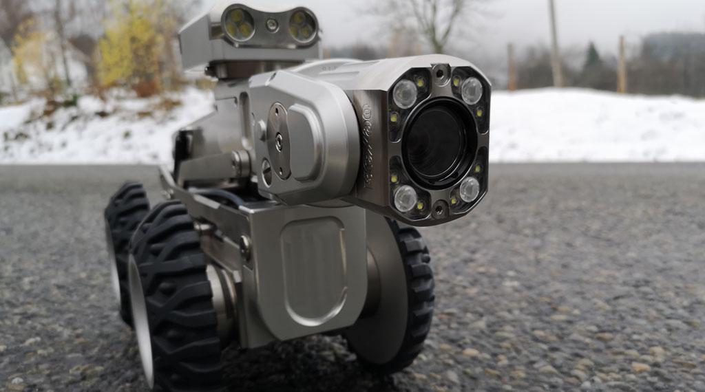 En robot med kamera og hjul står på en grå asfaltvei, omgitt av snø og trær i bakgrunnen.