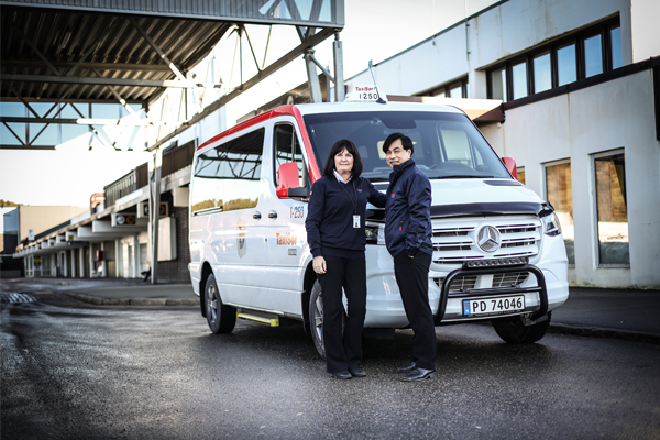 To personer står foran en hvit taxi med rød dekorasjon, parkert i et industriområde.