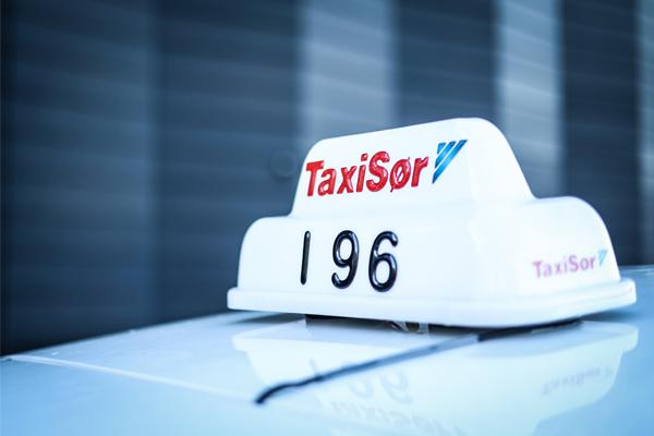 Takskilt fra TaxiSør med nummeret 196 på taket av en hvit taxi.
