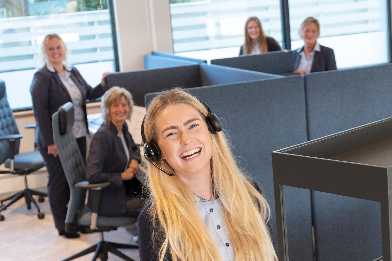 En smilende kvinne med headset står foran en kontorlandskap med flere kolleger i bakgrunnen.