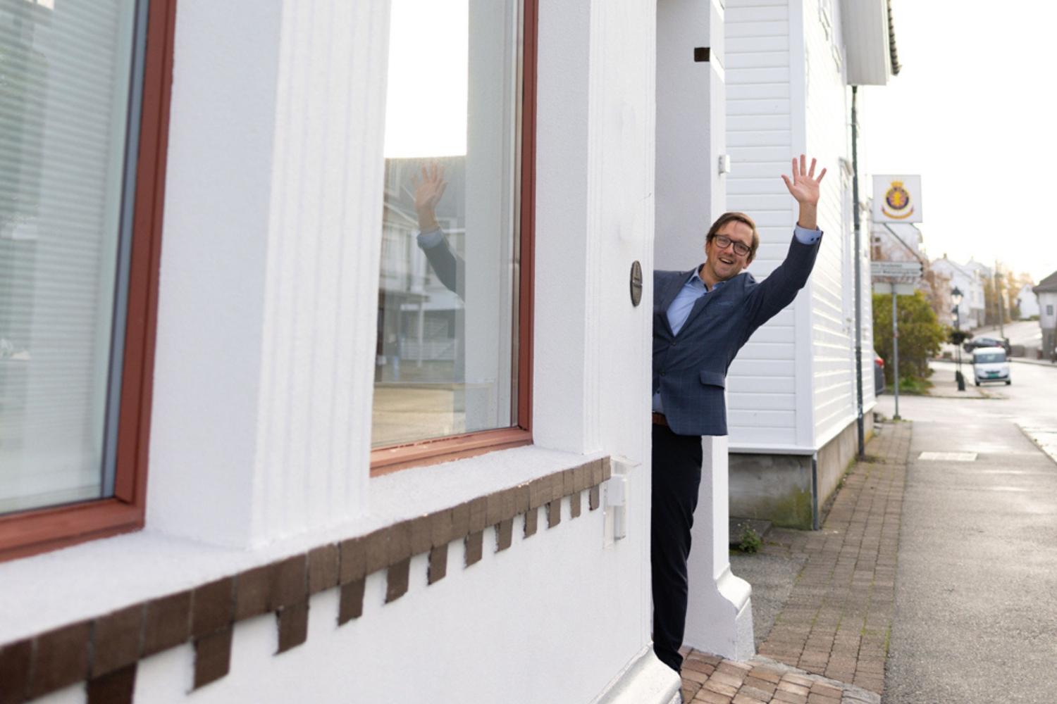 En mann i dress står i døren til et hvitt hus og vinker med hånden.
