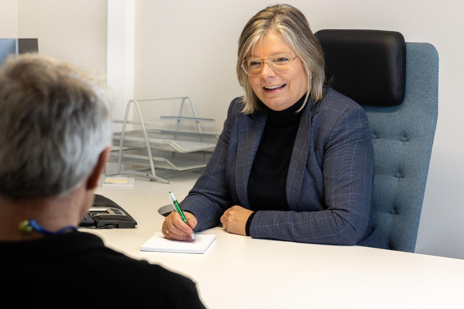 En kvinne i blå blazer sitter ved et skrivebord og smiler mens hun skriver notater. En mann sitter med ryggen til.