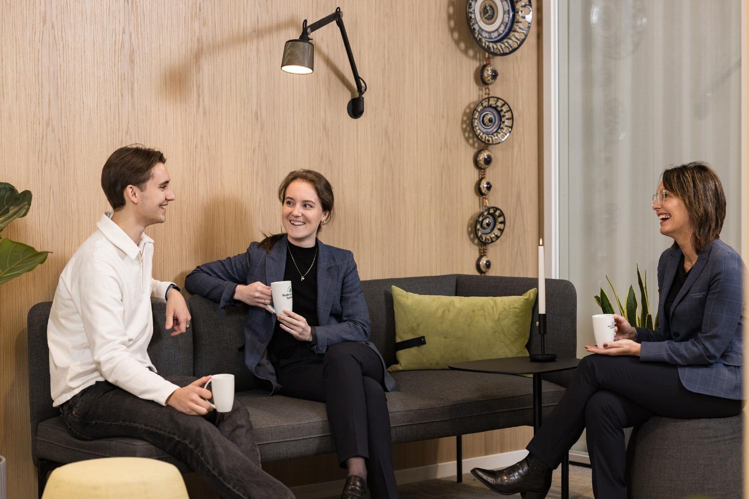 Tre personer sitter i en moderne stue, smilende og holder kopper, med planter og dekorative elementer i bakgrunnen.