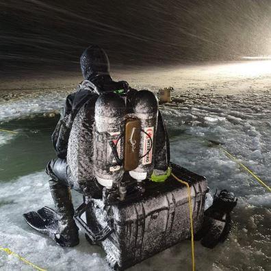 Dykker i vinterklær sitter på en boks ved isvann, omringet av snø og vind.