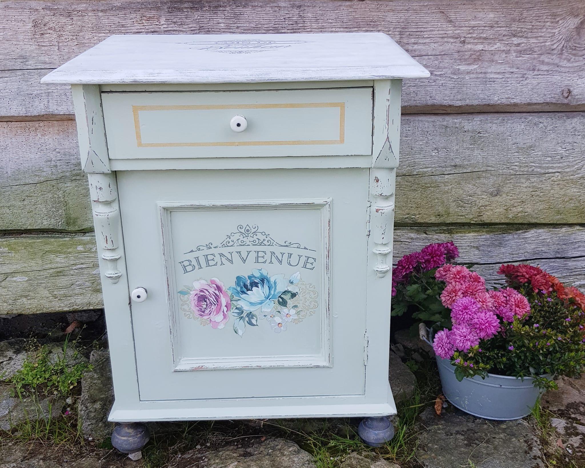 Grønn kommode med en skuff og dekorative blomster, plassert ved en vegg med blomsterpotte ved siden av.