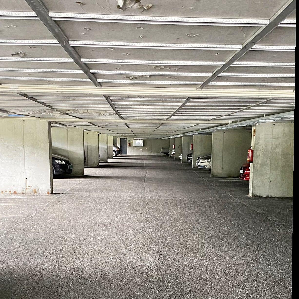 En tom parkeringskjeller med flere parkerte biler og betongvegger. Belysningen er dempet og taket er grått.