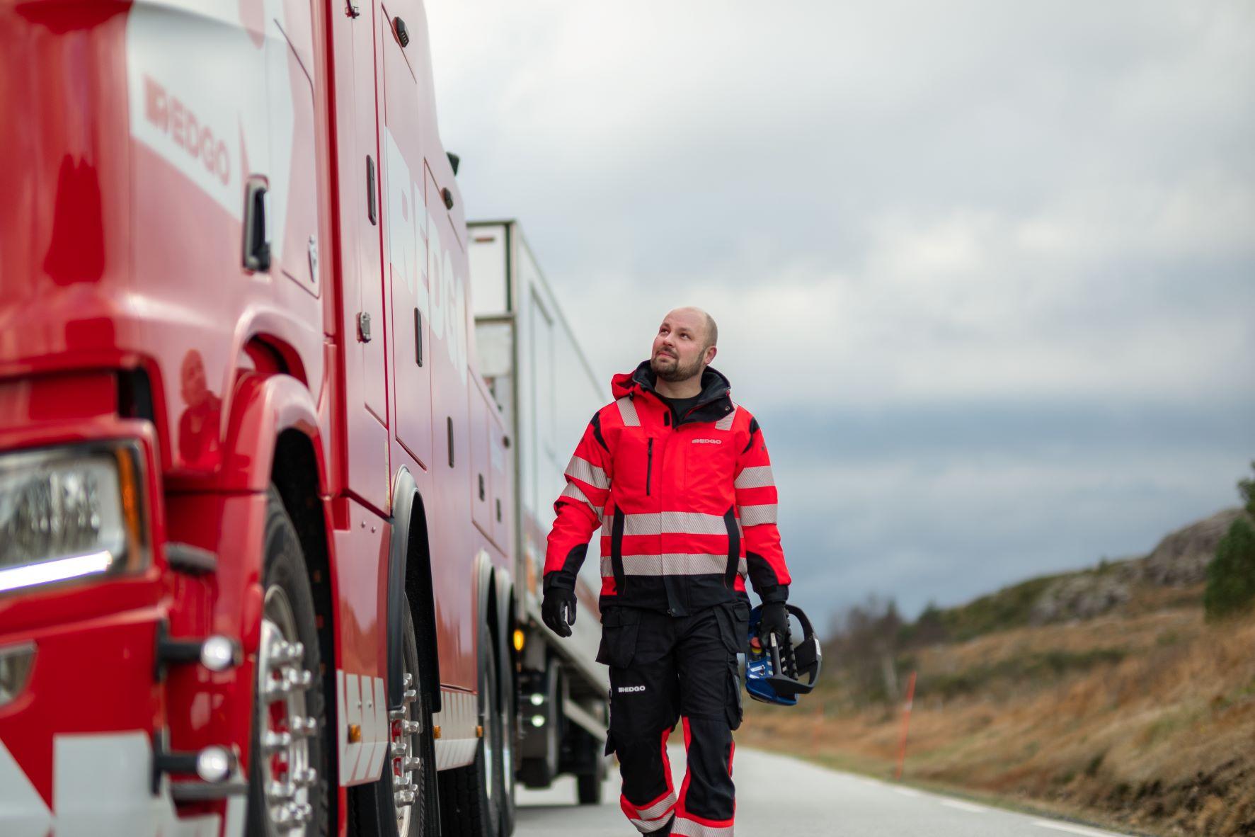 En redningsmann i rød uniform ser opp mot en stor redningsbil langs veien.