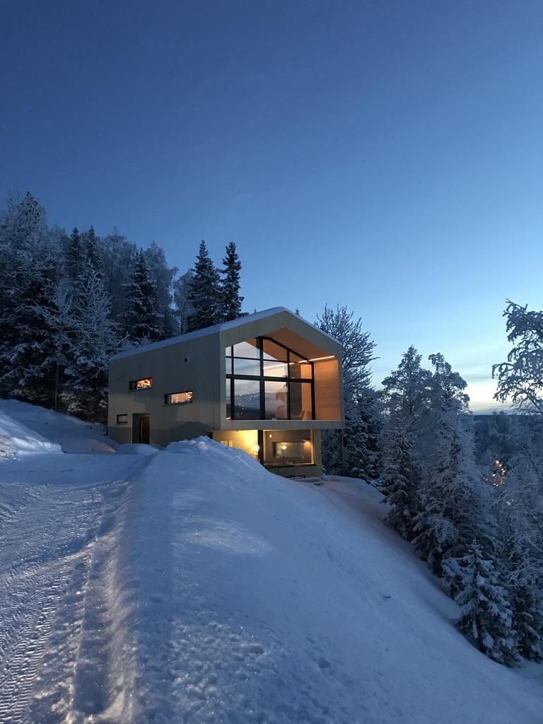 Moderne hytte i snødekt landskap med store vinduer og lysende interiør, omgitt av trær i skumringen.