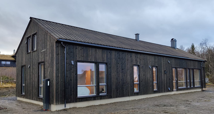 Moderne, svart trehus med store vinduer og skråtak, beliggende i et naturskjønt område.