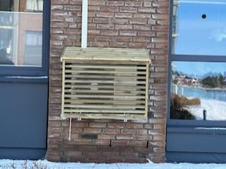 Klimaanlegg med trebeskyttelse montert på murvegg ved vindu. Snø på bakken og utsikt til vann i bakgrunnen.