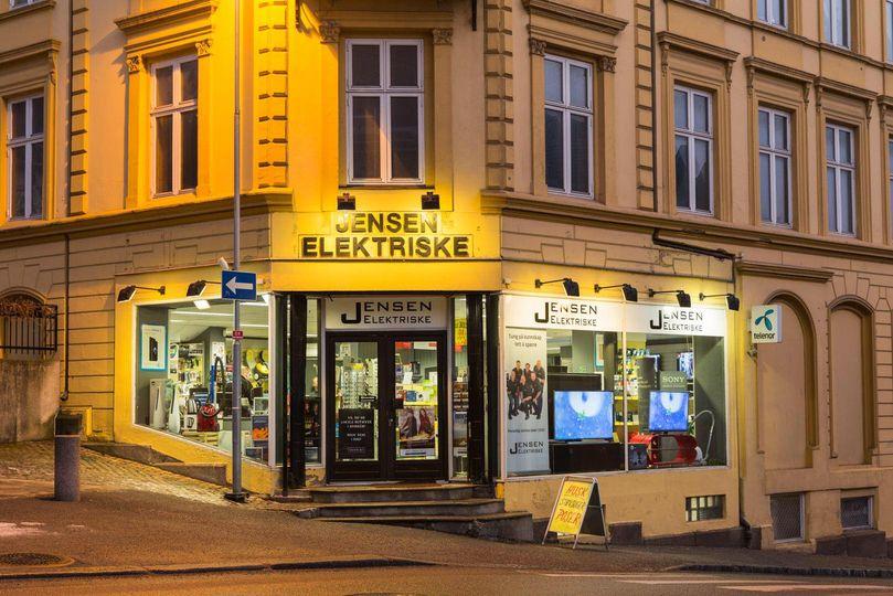Butikkfasade med navnet "Jensen Elektriske" opplyst om kvelden, med vinduer som viser elektronikkprodukter.