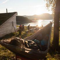 En person ligger avslappet i en hengekøye med solnedgang over vannet og folk som samles i bakgrunnen.