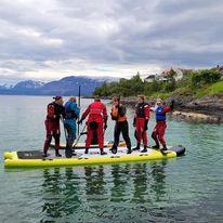 Gruppe på en stor paddleboard i fjorden, iført våtdrakter, med fjell i bakgrunnen.