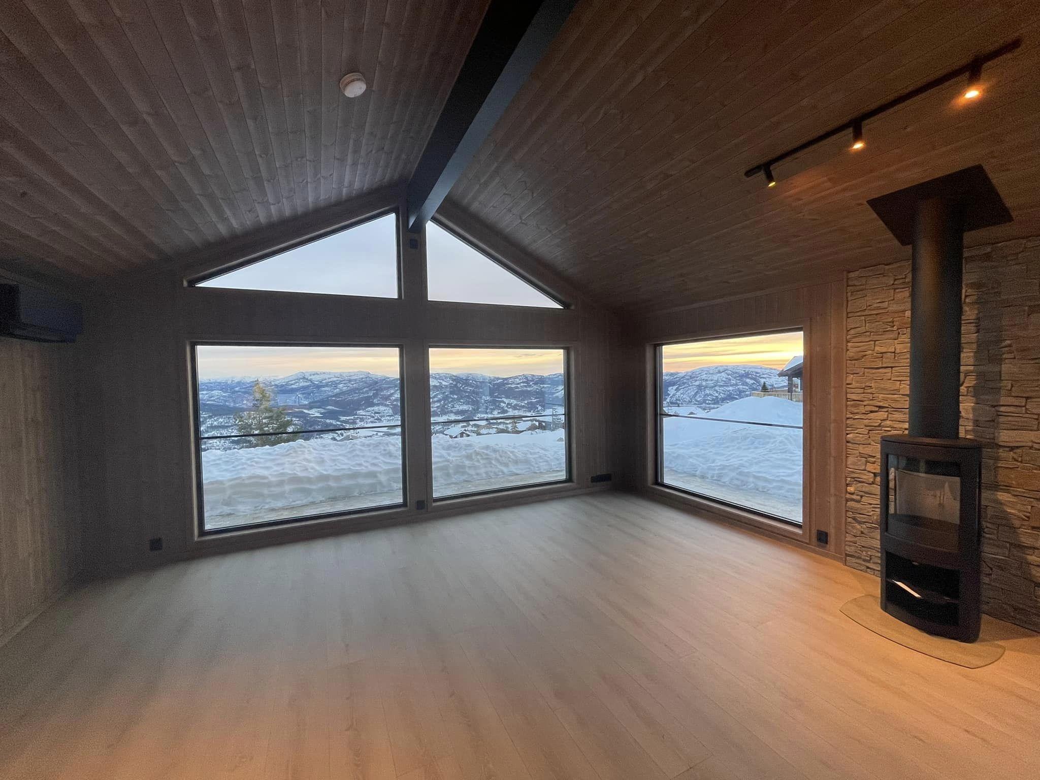 Moderne stue med store vinduer som gir utsikt over snødekte fjell og en peis i hjørnet.