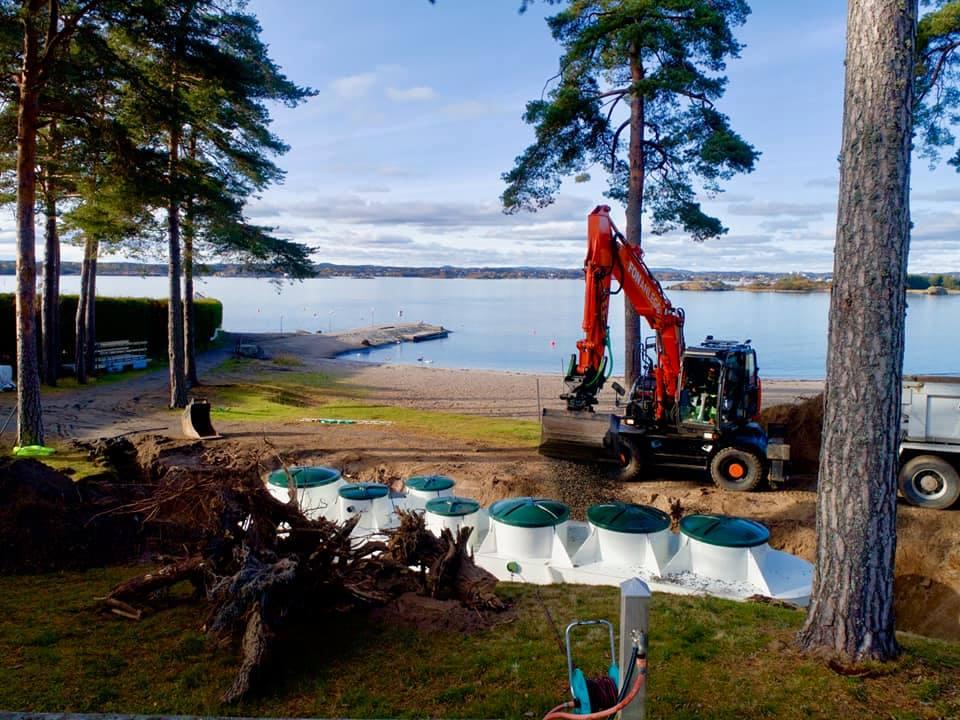Gravemaskin arbeider ved stranden, med trær og en båt i bakgrunnen, på en solfylt dag.