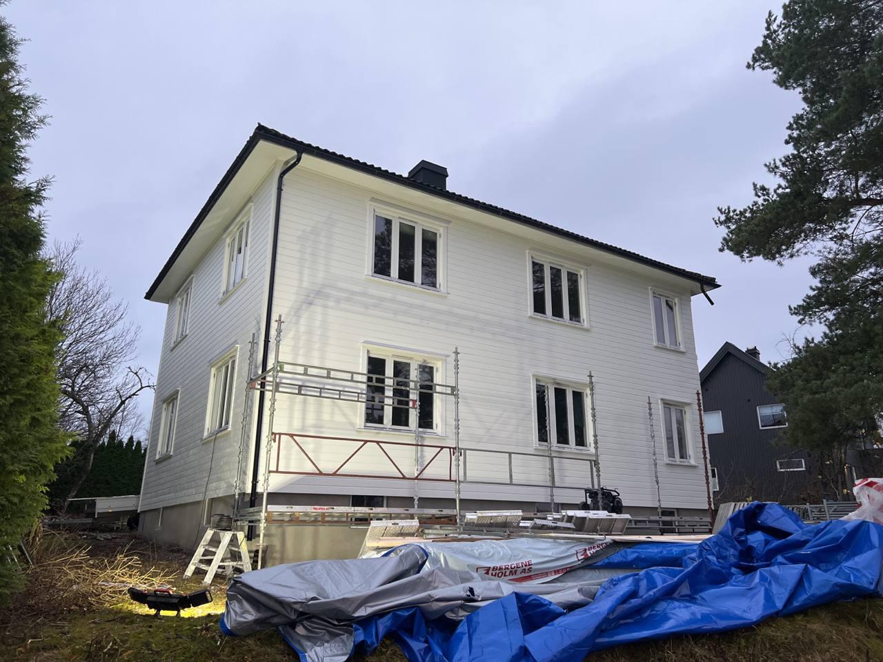 Et hvitt hus med stillas og blå presenning i hagen, omringet av trær og en grå bygning i bakgrunnen.