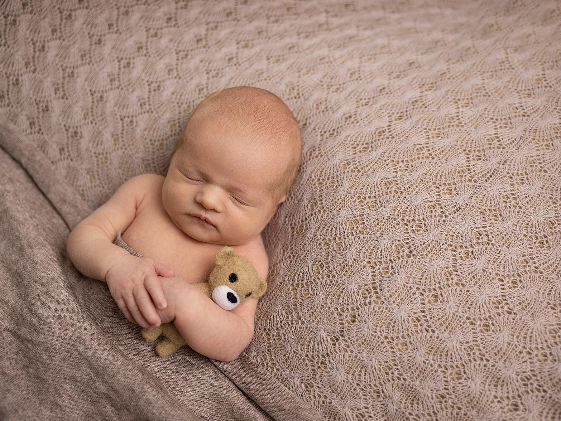 Nyfødt baby som sover på en beige strikket teppe, holder en liten kosedyrbjørn i armene.