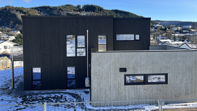 Moderne hus med svart og grå fasade, omgitt av snø og landskap med fjell i bakgrunnen.