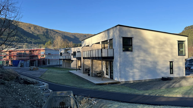 Moderne bygning med balkonger, omgitt av fjell og byggearbeid i bakgrunnen, under klar blå himmel.