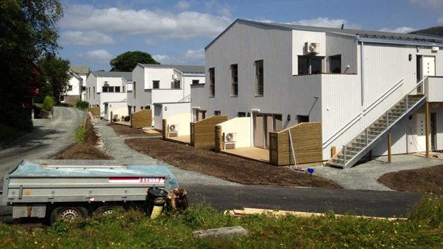 Moderne boliger med trehus og trapper, beliggende langs en grusvei med en tilhenger i forgrunnen.