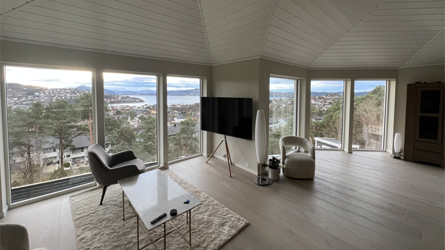 Moderne stue med store vinduer, TV, sofaer og utsikt over landskapet og vannet.