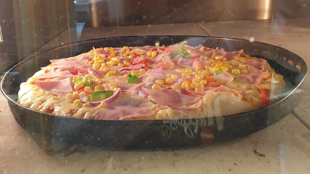 En nybakt pizza med skinke, paprika, løk og mais i en svart stekepanne.