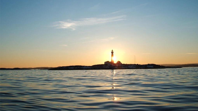 En fyrtårn på en øy ved solnedgang, med rolig hav og klar himmel i bakgrunnen.