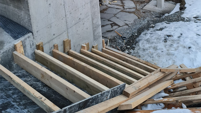 Bilde av en byggeplass med treplanker stablet på bakken ved en betongvegg og snø i bakgrunnen.